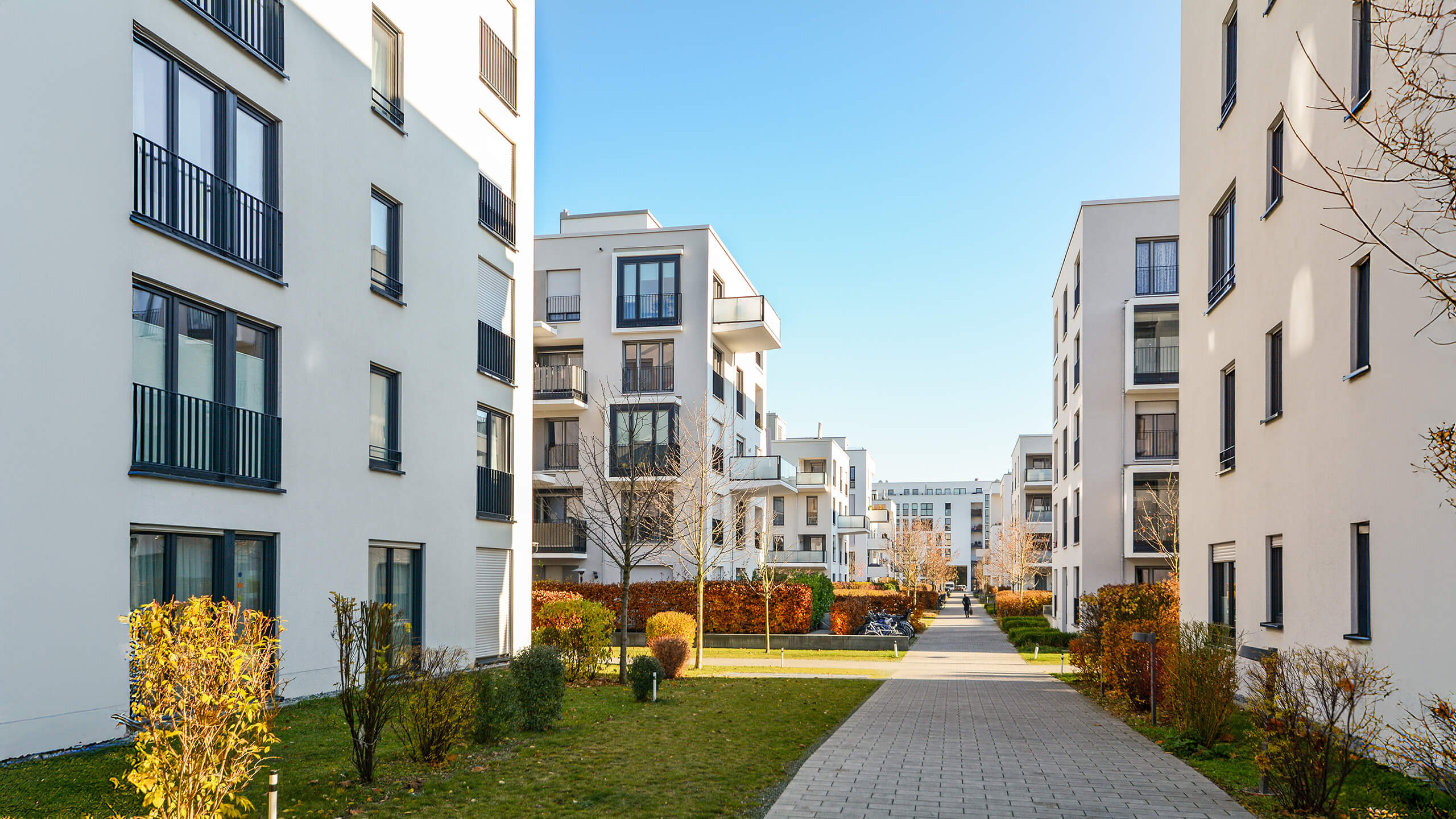 Wohnblöcke mit Balkonen an Weg, umgeben von Grün und blauem Himmel.
