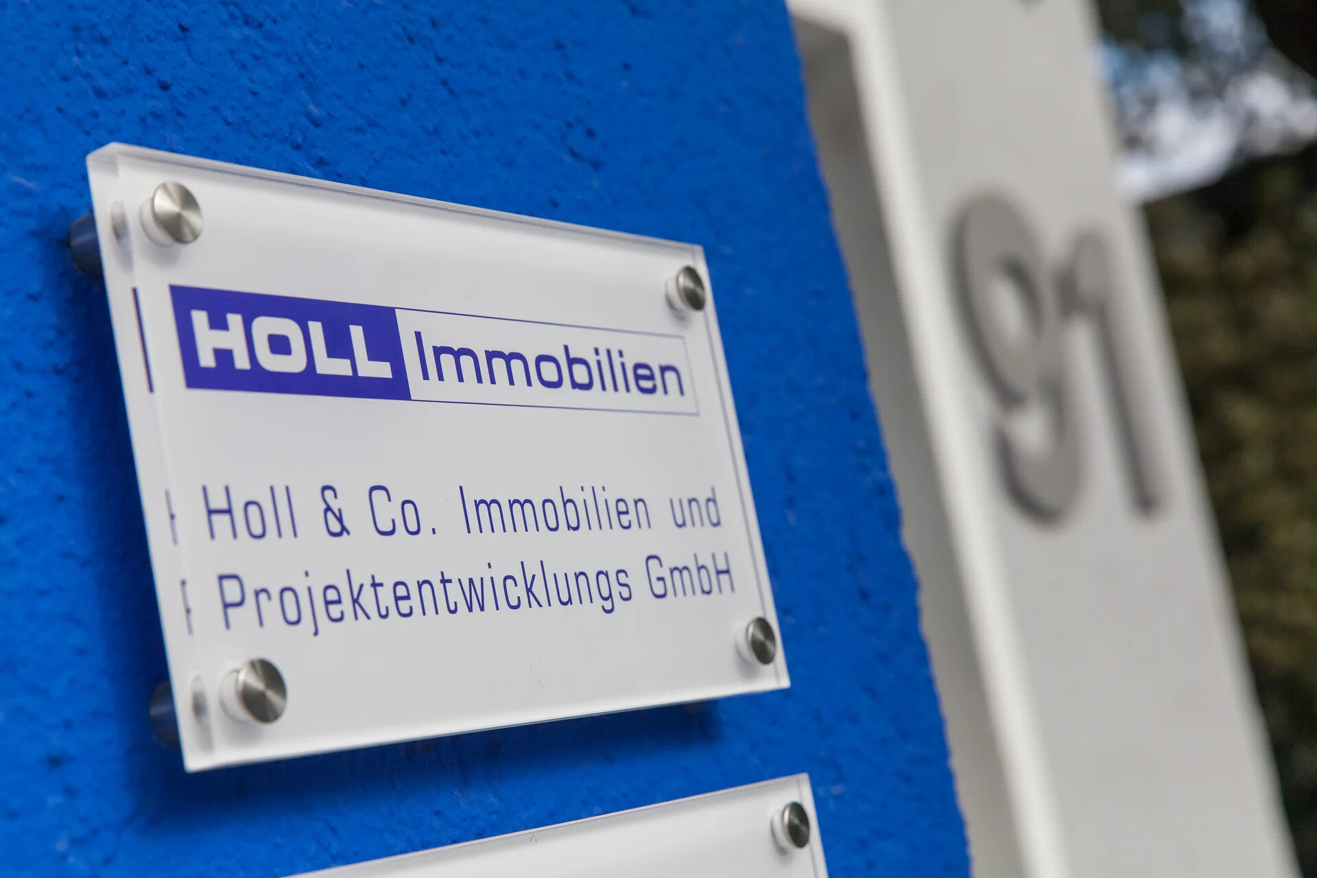 Firmenschild von HOLL Immobilien an einer blauen Außenwand.