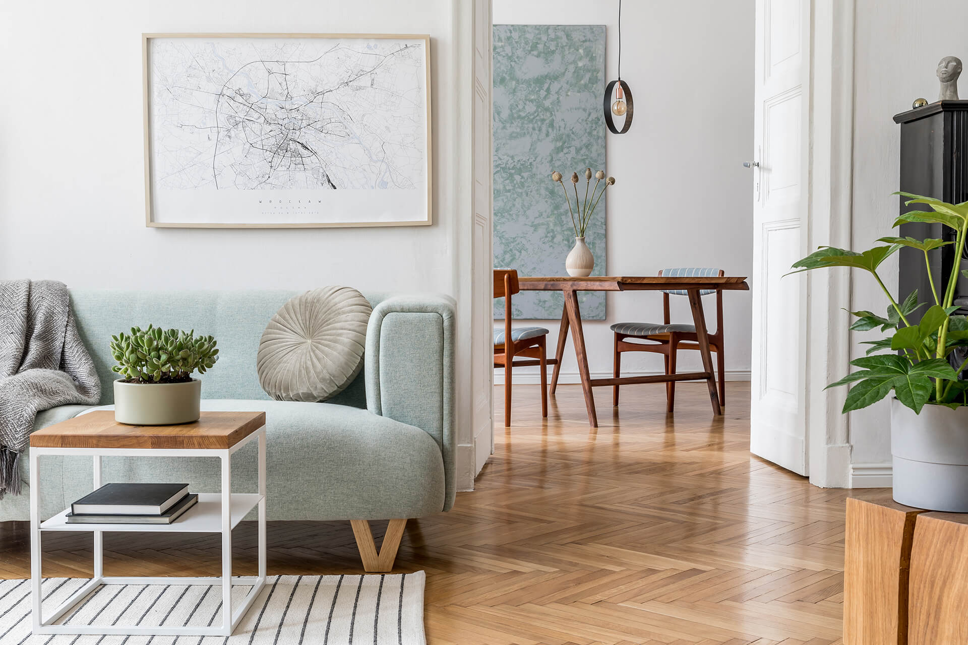 Helles Wohnzimmer mit hellblauem Sofa, kleinem Beistelltisch mit Pflanze, Wandbild einer Stadtkarte und Blick in den Essbereich mit Holztisch und Stühlen