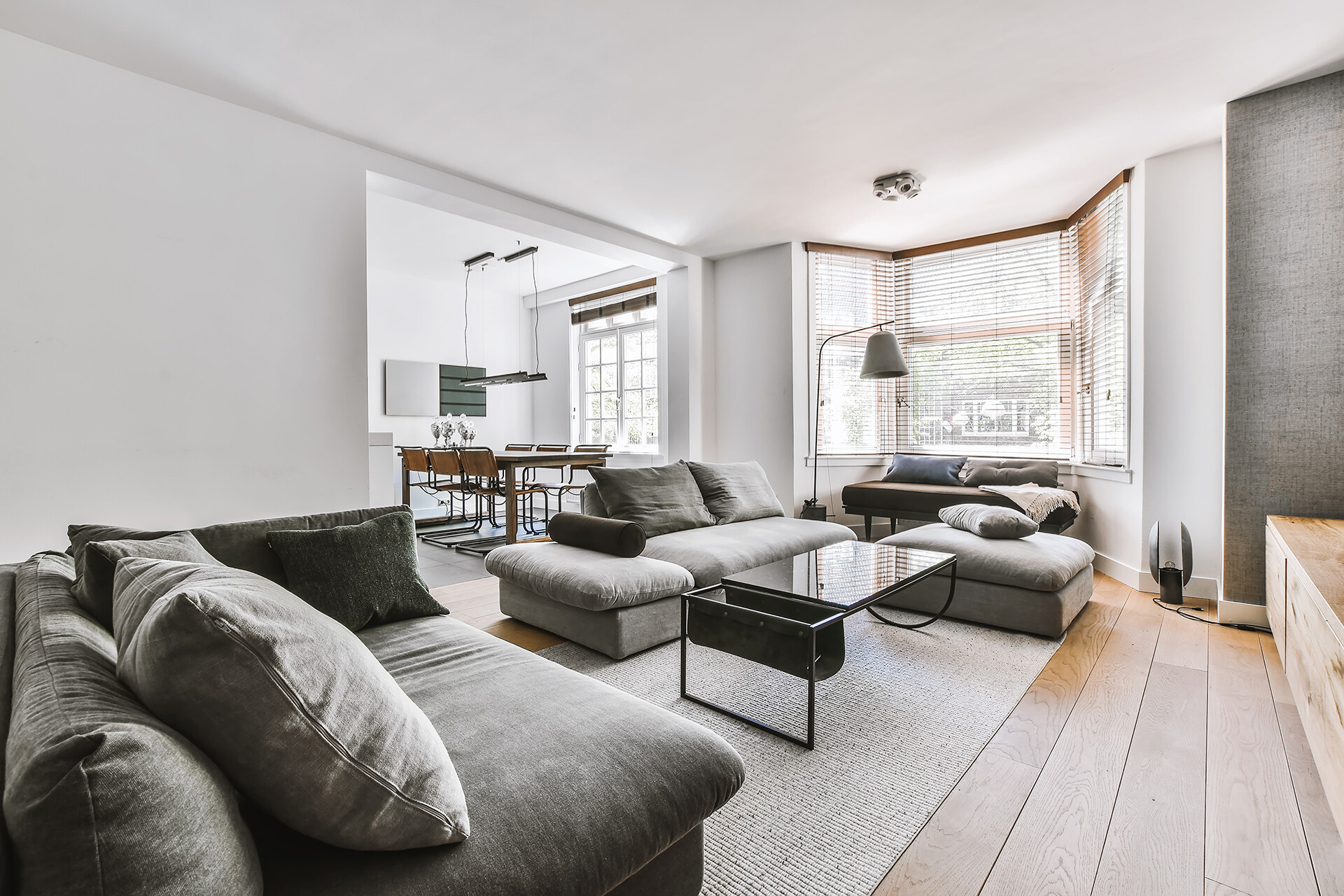 Helles Wohnzimmer mit mehreren grauen Sofas, einem schwarzen Couchtisch auf einem Teppich und großen Fenstern mit Holzjalousien