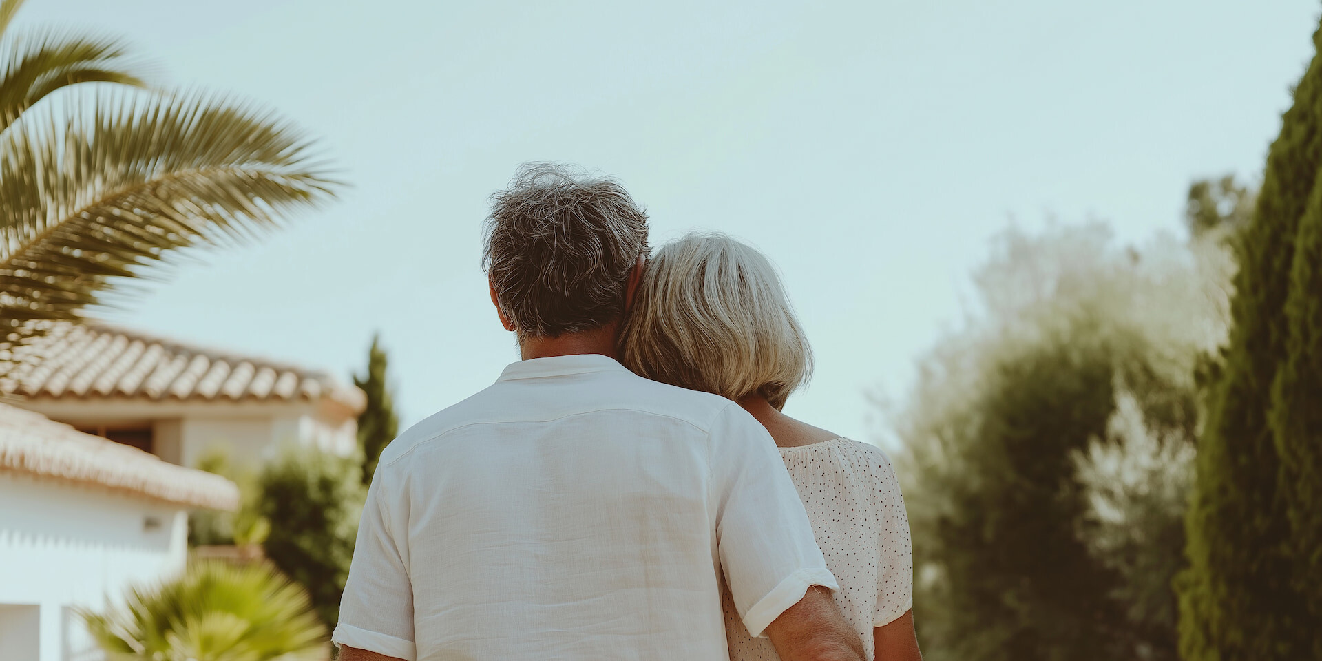 Älteres Paar umarmt sich draußen vor Palmen und Haus