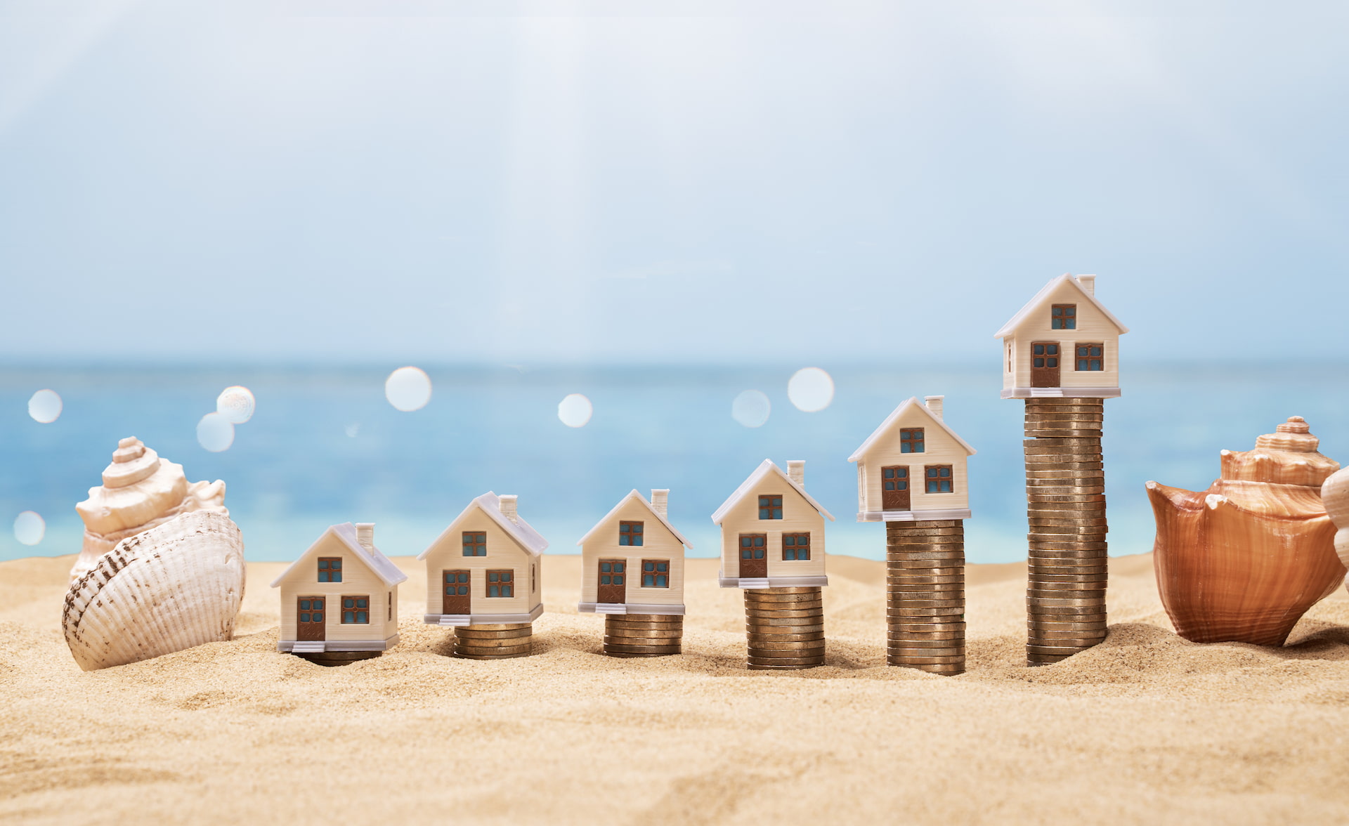 Hausmodelle auf Münzen im Sand, Meer und Muscheln im Hintergrund