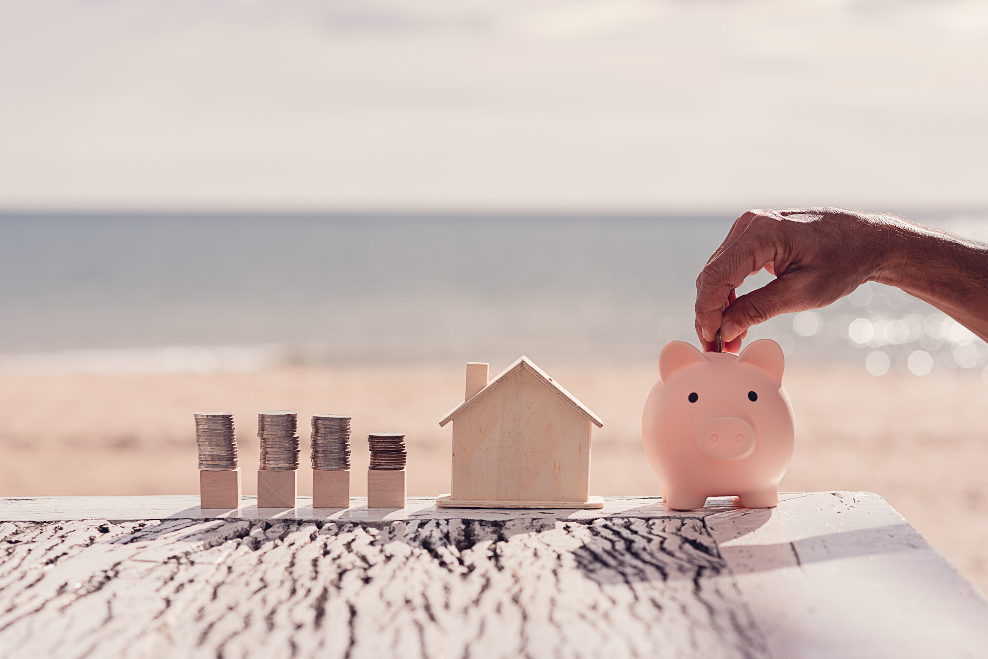 Hand legt Münze in Sparschwein neben Hausmodell und Münzstapel am Strand