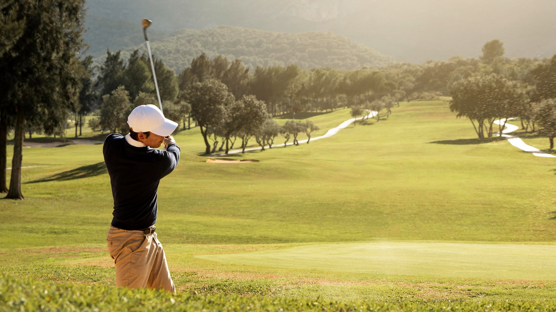 Golfer mit weißer Mütze schwingt den Schläger auf einem sonnigen, grünen Platz