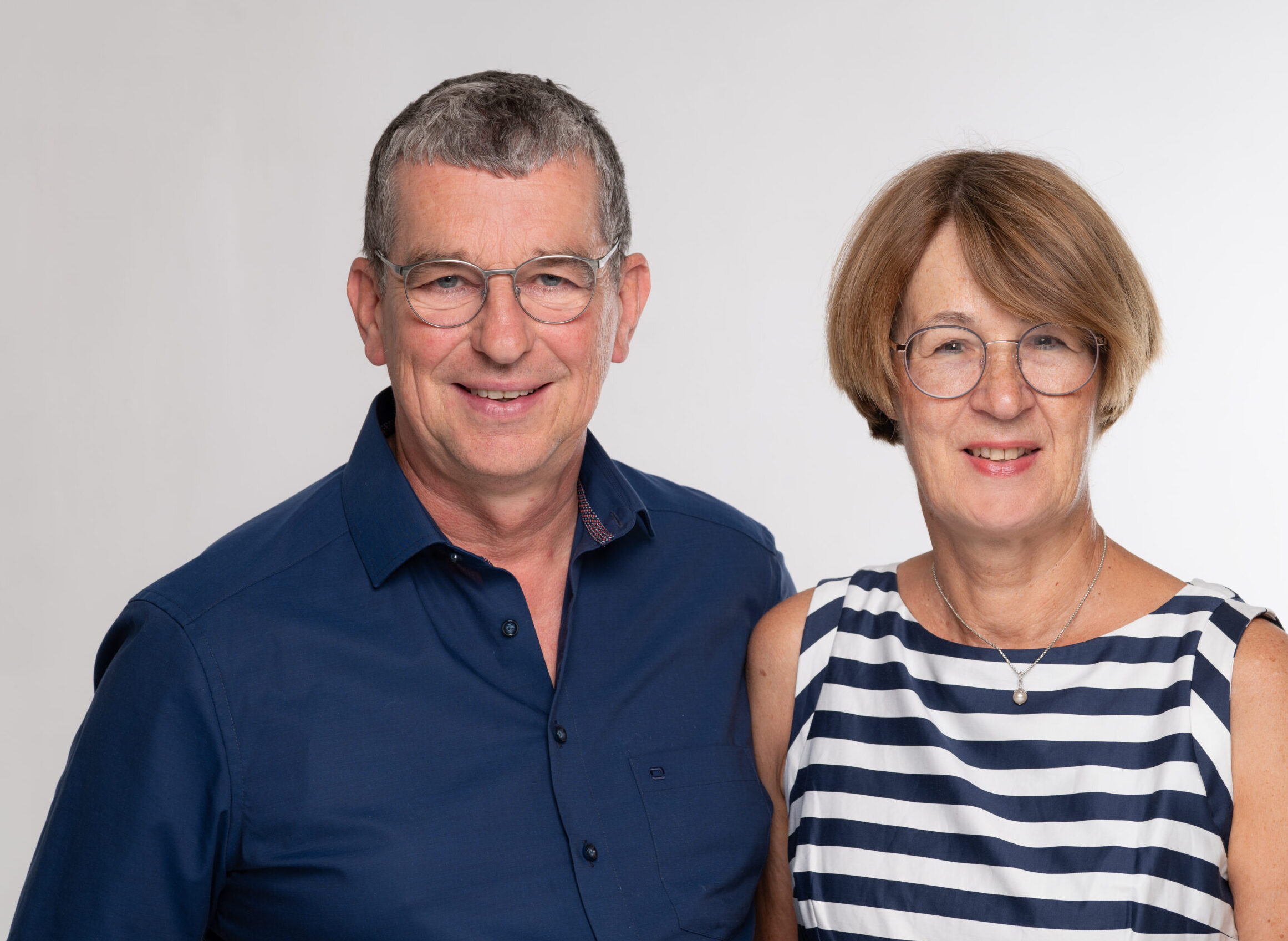 Ein freundliches Porträt von Olaf Hübner und Diana Rutel nebeneinander vor einem neutralen, hellgrauen Hintergrund. Olaf Hübner trägt ein dunkelblaues Hemd und eine Brille, Diana Rutel trägt ein blau-weiß gestreiftes ärmelloses Oberteil, eine Brille und eine dezente Kette mit Perlenanhänger. Beide blicken lächelnd direkt in die Kamera.
