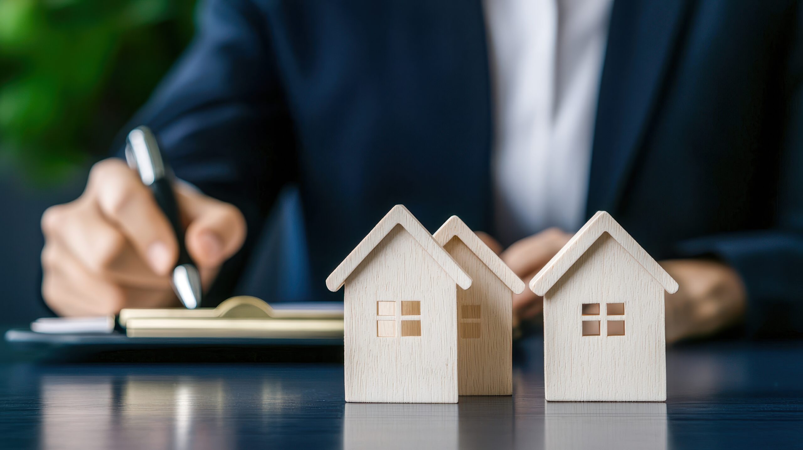 Drei hölzerne Modellhäuser stehen im Vordergrund, im Hintergrund sitzt eine Person und schreibt etwas auf