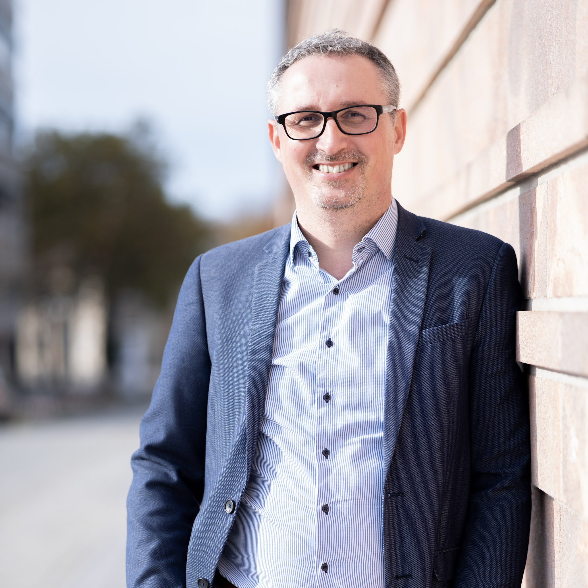 Ein lächelnder Immobilienmakler Andreas Langer mit Brille im Business-Anzug steht draußen an einer Steinwand und blickt direkt in die Kamera.