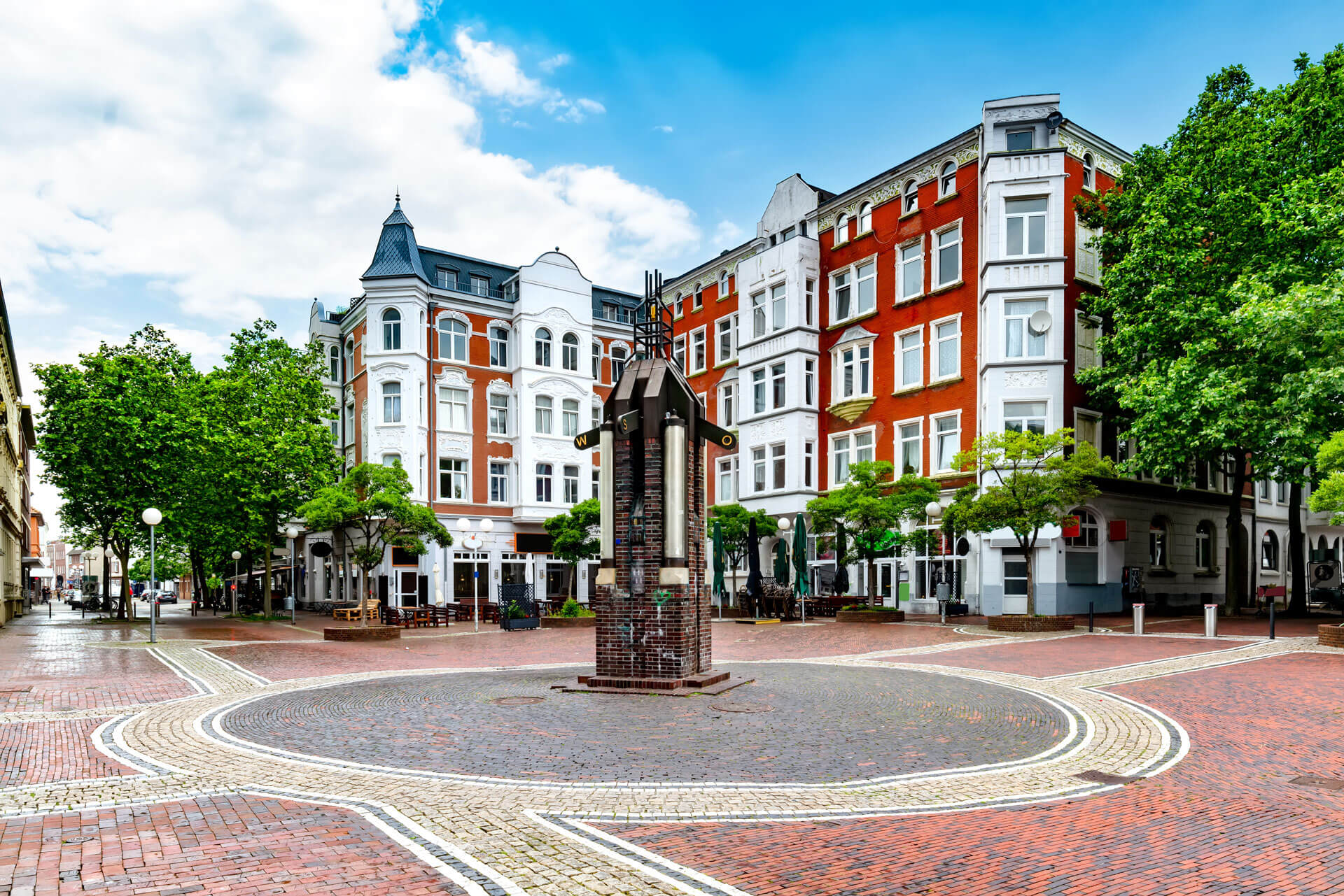 Ein Platz aus Backstein, ein kleines Denkmal, historische rote und weiße Gebäude, Bäume.
