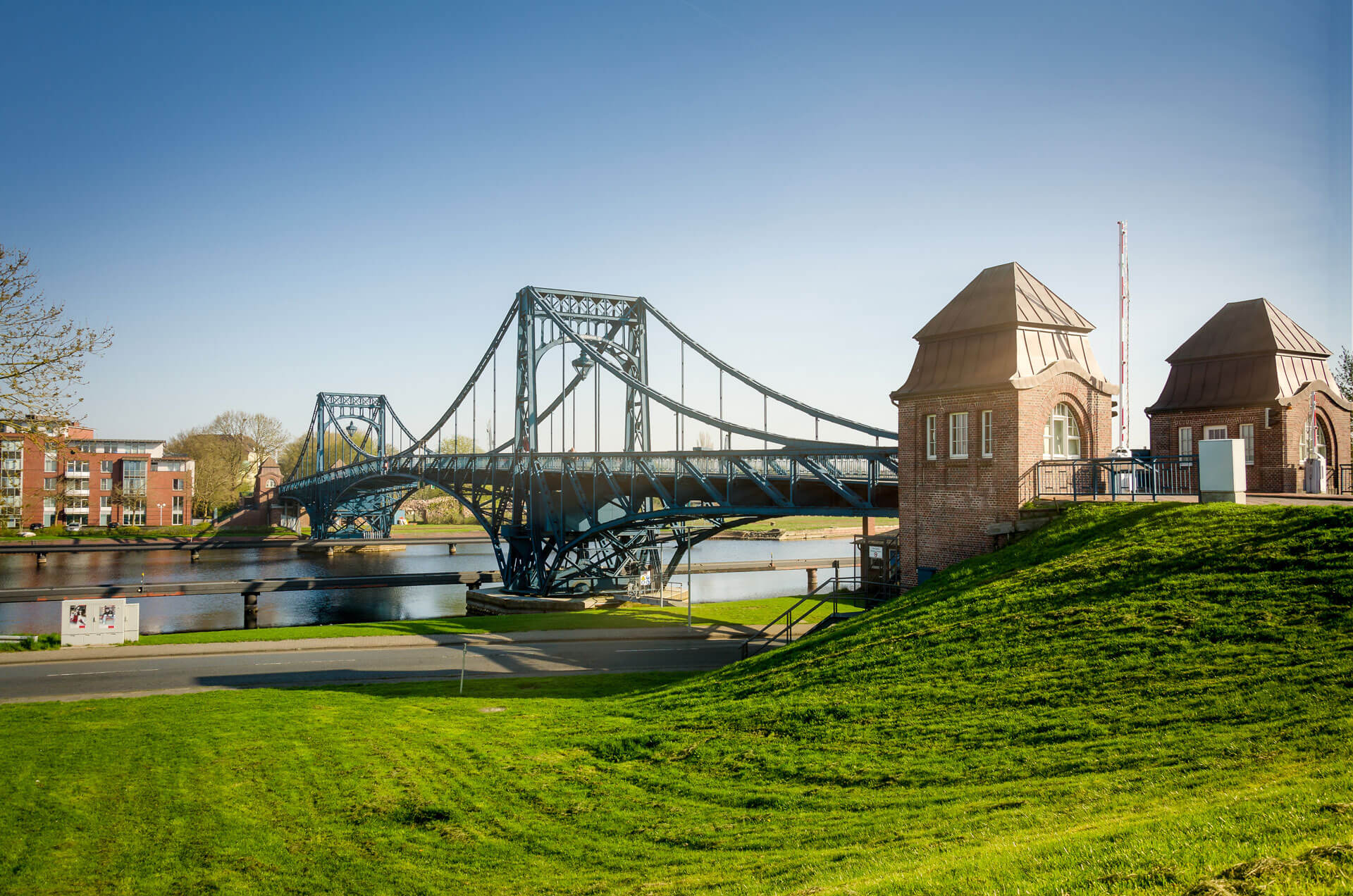 Blaue Eisenbrücke mit Steintürmen überquert Fluss, grüne Wiese.