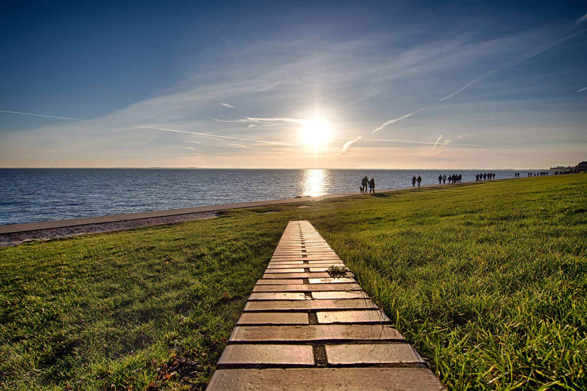 Sonnenuntergang am Meer, Leute spazieren auf grasigem Uferweg.
