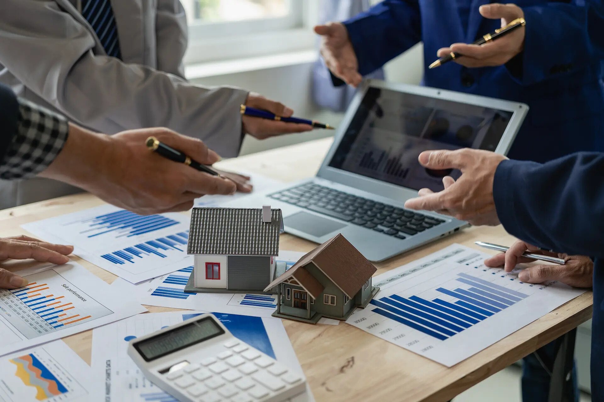 Vier Personen mit Finanzunterlagen, Laptop und zwei Hausmodellen am Tisch.