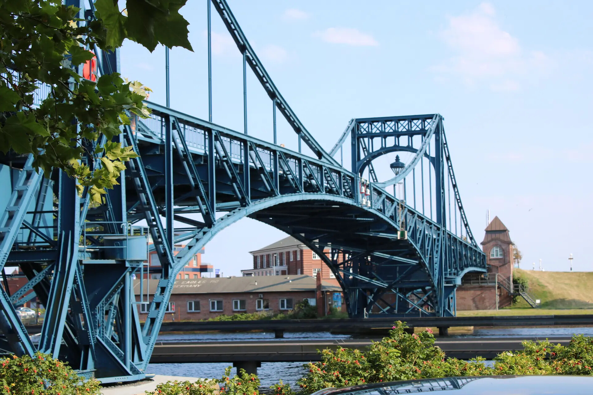 Blaue Stahlbrücke mit Bogen über Wasser, Gebäude im Hintergrund.
