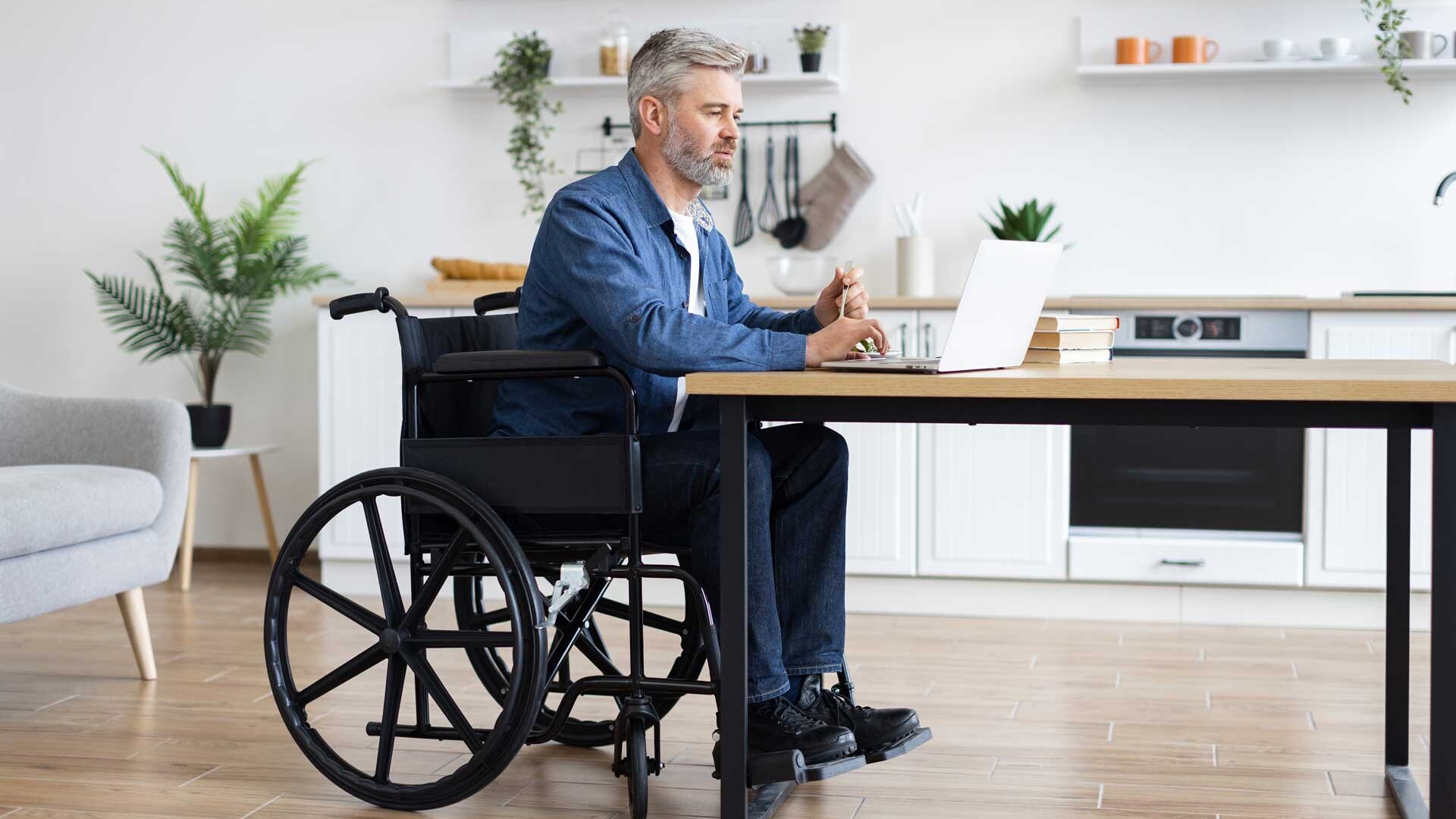 Mann im Rollstuhl sitzt mit Laptop am Küchentisch in seiner barrierefreien Wohnung