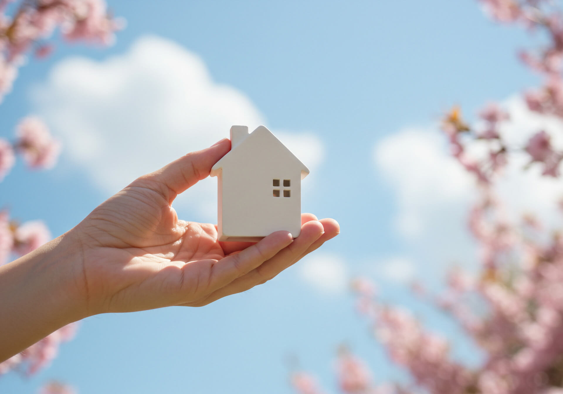 Hand hält weißes Hausmodell, draußen mit Blumen und blauem Himmel