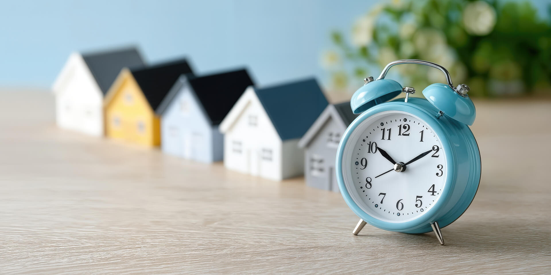 Blauer Wecker auf Tisch vor Modellhäusern und Blumen.