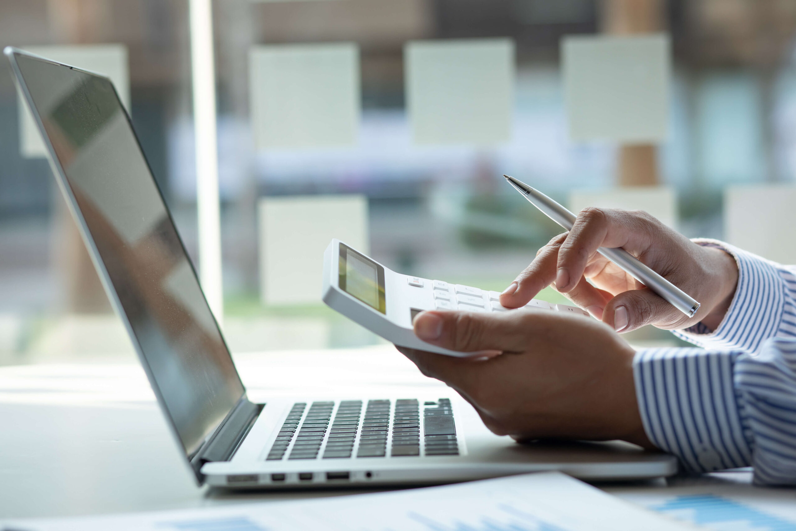 Person kalkuliert am Laptop mit Stift und Dokumenten auf dem Tisch