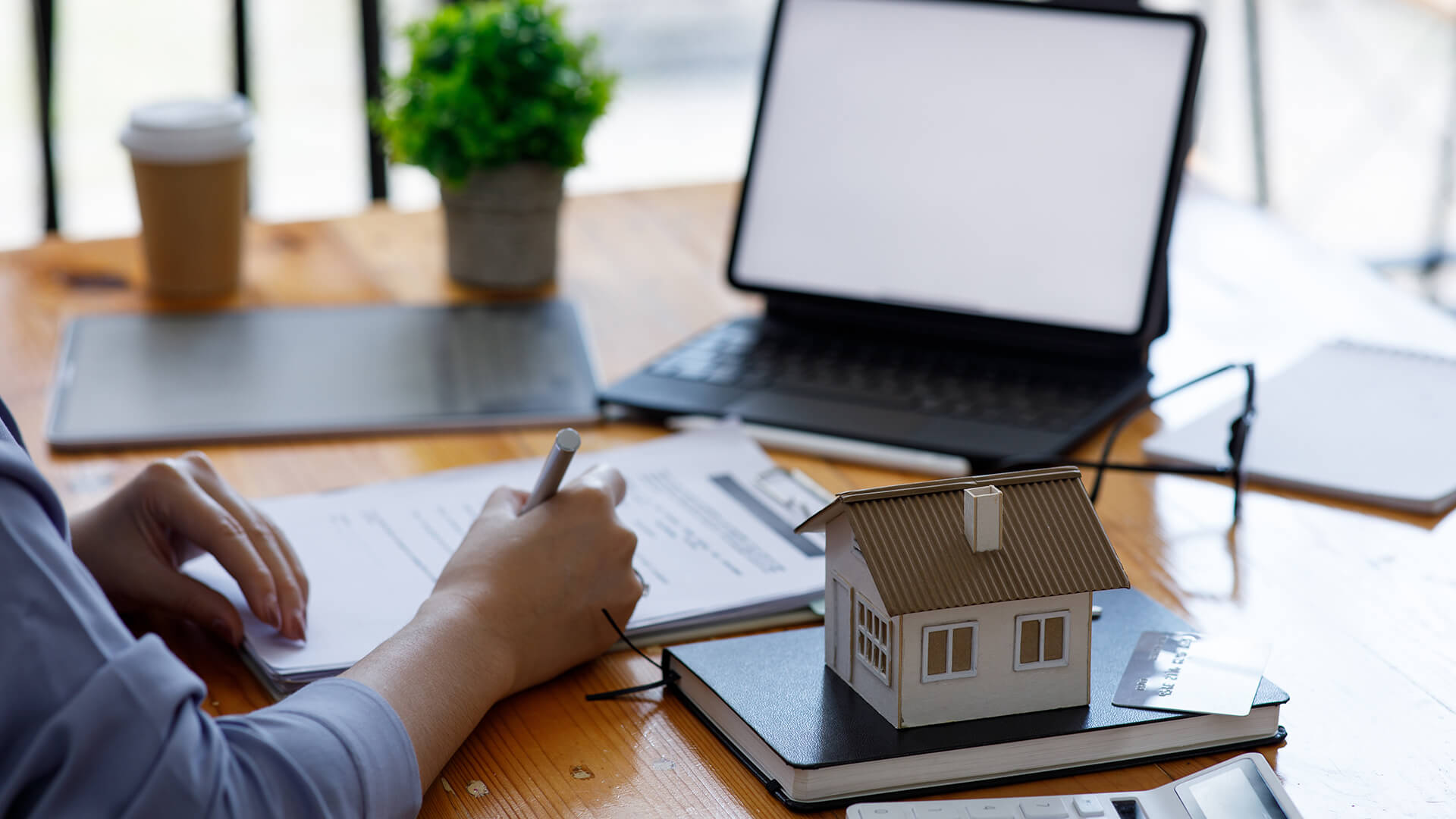 Person schreibt an Schreibtisch mit Laptop und Modellhaus