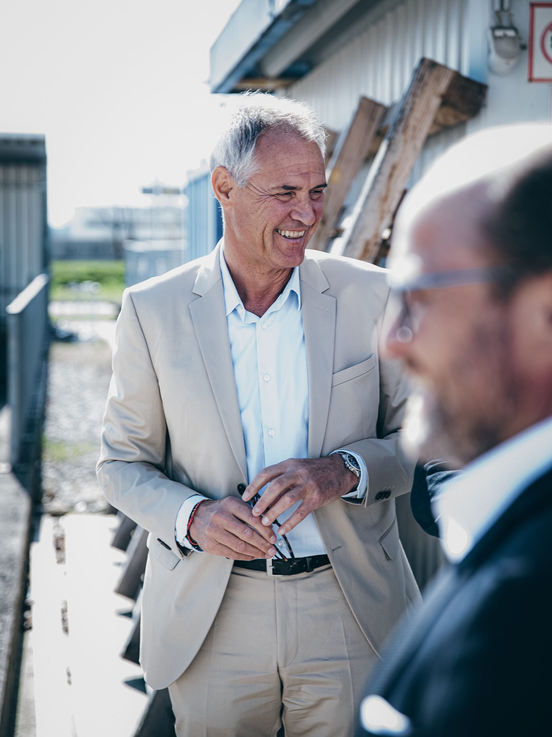 Mann im beigen Anzug steht draußen, vorne weitere Person.