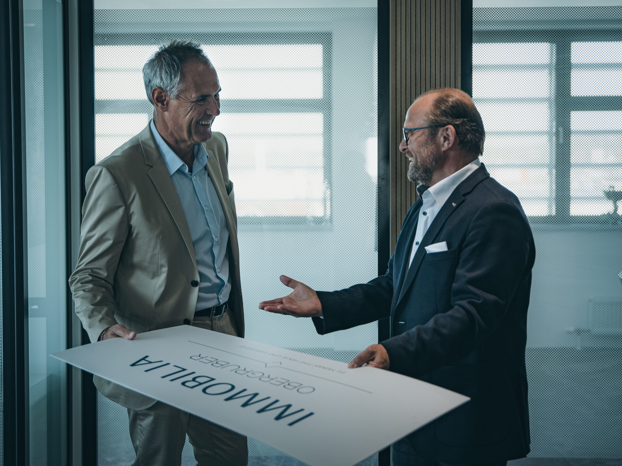 Zwei Männer im Büro halten lächelnd ein IMMOBILIA-Schild