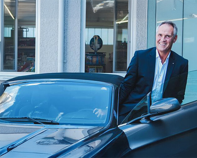 Mann im Anzug lächelt neben geöffneter Autotür vor Gebäude