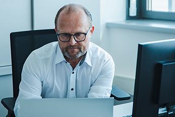 Mann mit Brille arbeitet am Schreibtisch mit Laptop und Monitor