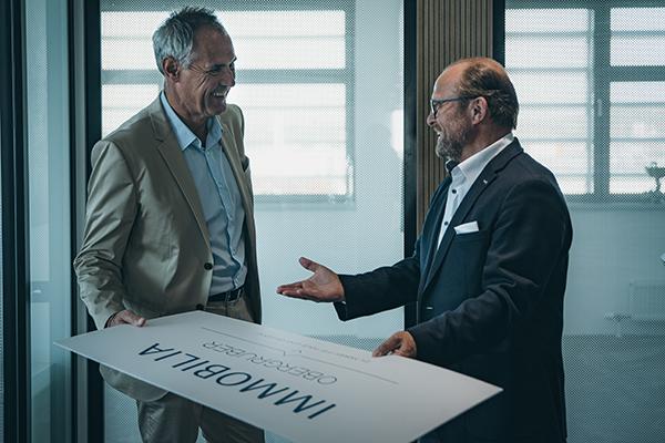Zwei Männer im Büro halten lächelnd ein IMMOBILIA-Schild