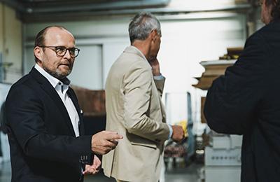 Drei Männer im Anzug sprechen in Industrieumgebung, einer schaut zur Kamera