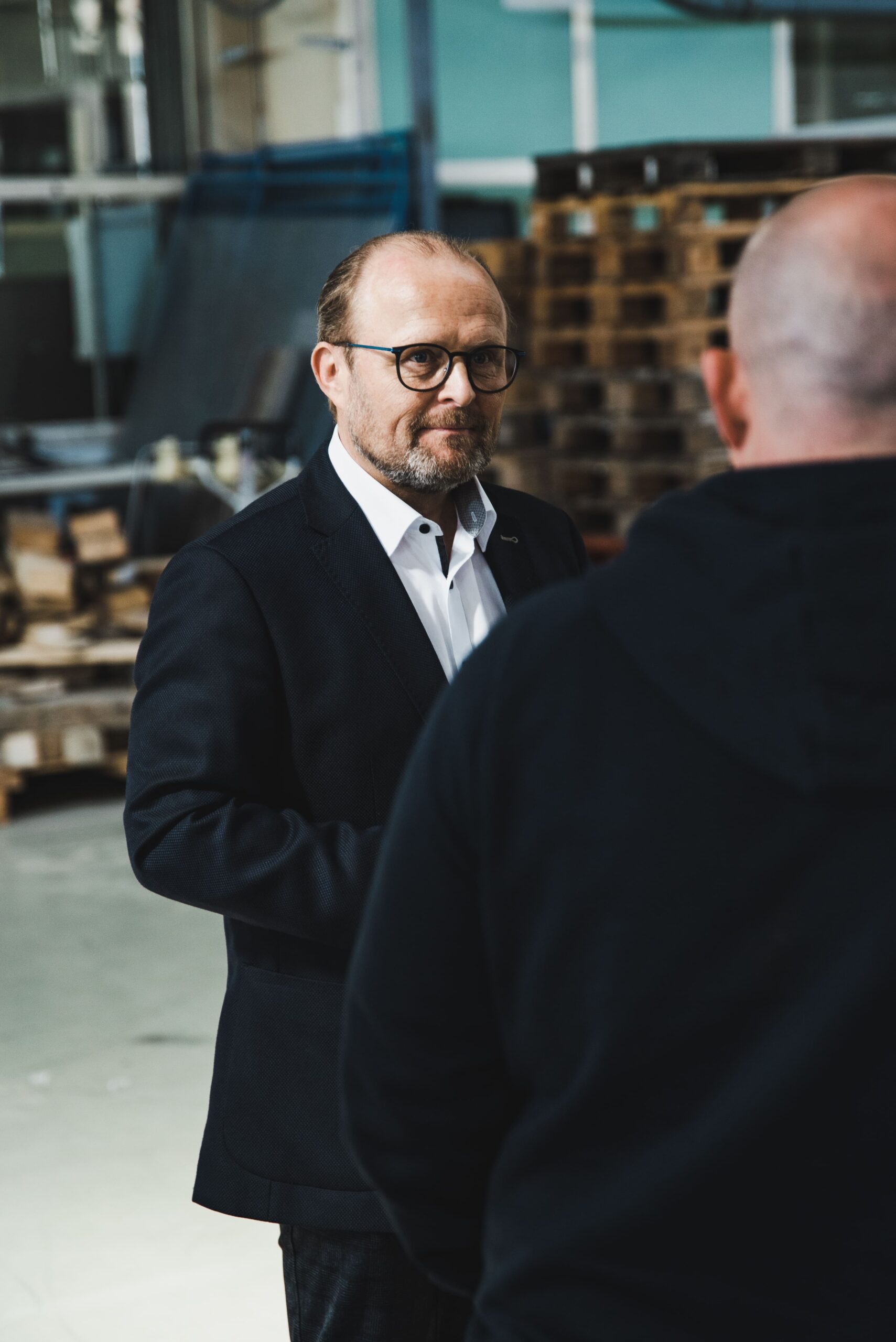 Mann mit Brille und Anzug spricht in einer Lagerhalle mit Person