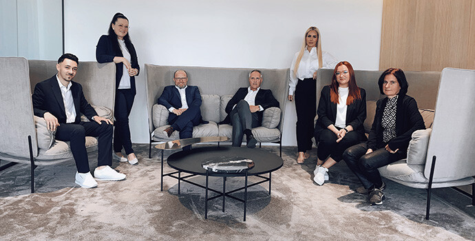 Sieben Personen im Anzug in moderner, neutraler Bürolounge.