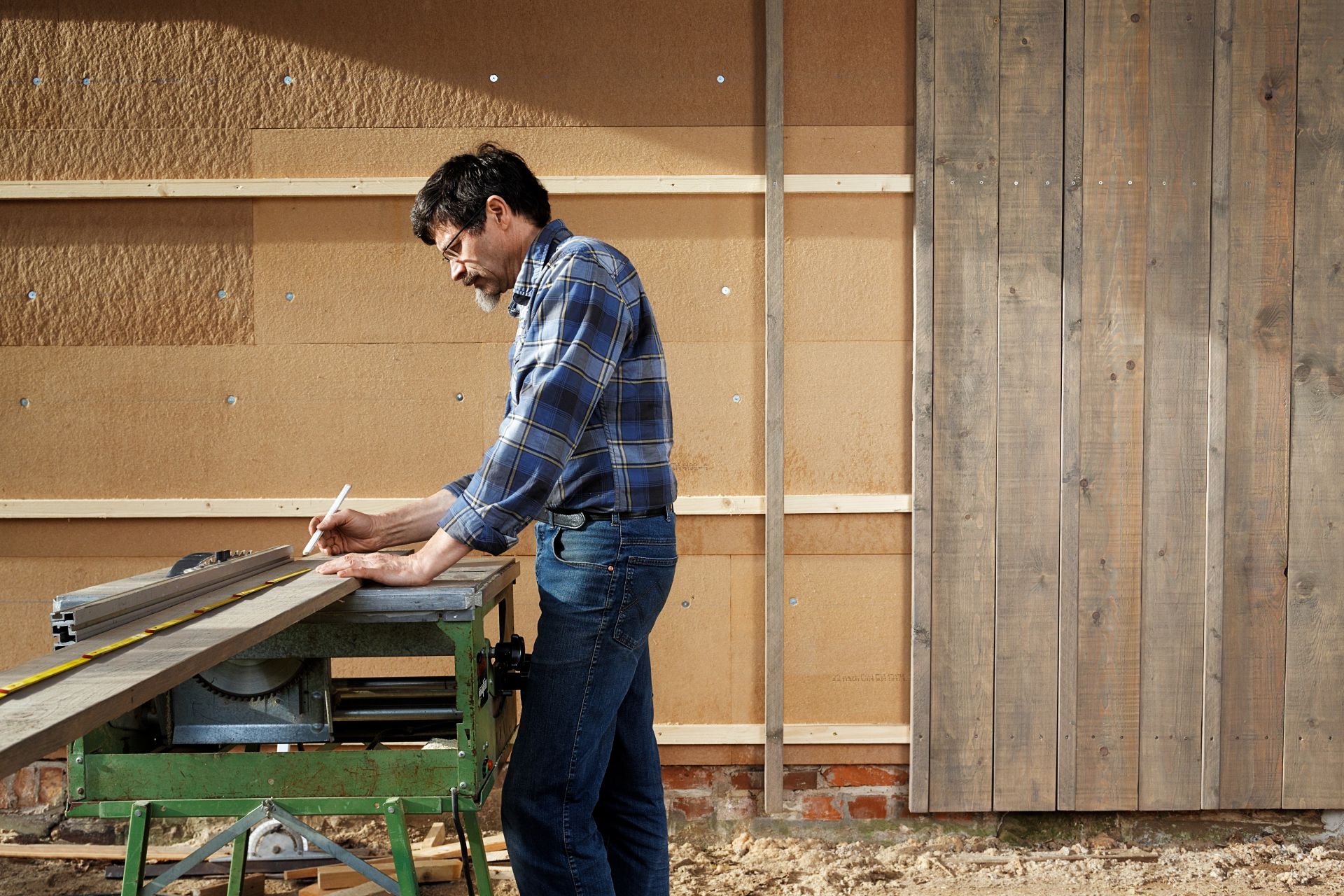 Mann misst und markiert Holz an Werkbank im Freien.