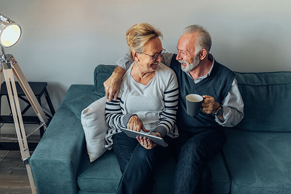 Älteres Paar auf dem Sofa trinkt Kaffee und schaut aufs Tablet.