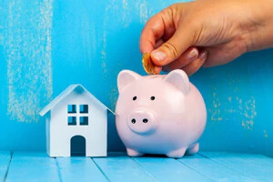 Eine Hand legt eine Münze in ein rosa Sparschwein bei einem weißen Haus auf einem blauen Tisch.