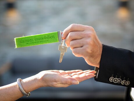 Eine Hand übergibt einen Schlüsselbund mit einem grünen Filzanhänger, auf dem "claussen immobilien IHR NORDHEIDE SPEZIALIST" steht, an eine andere Hand.