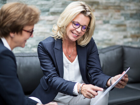 Zwei Frauen in Businesskleidung sitzen zusammen und schauen auf ein Dokument. Die Frau im Vordergrund mit blonden Haaren und Brille lächelt.