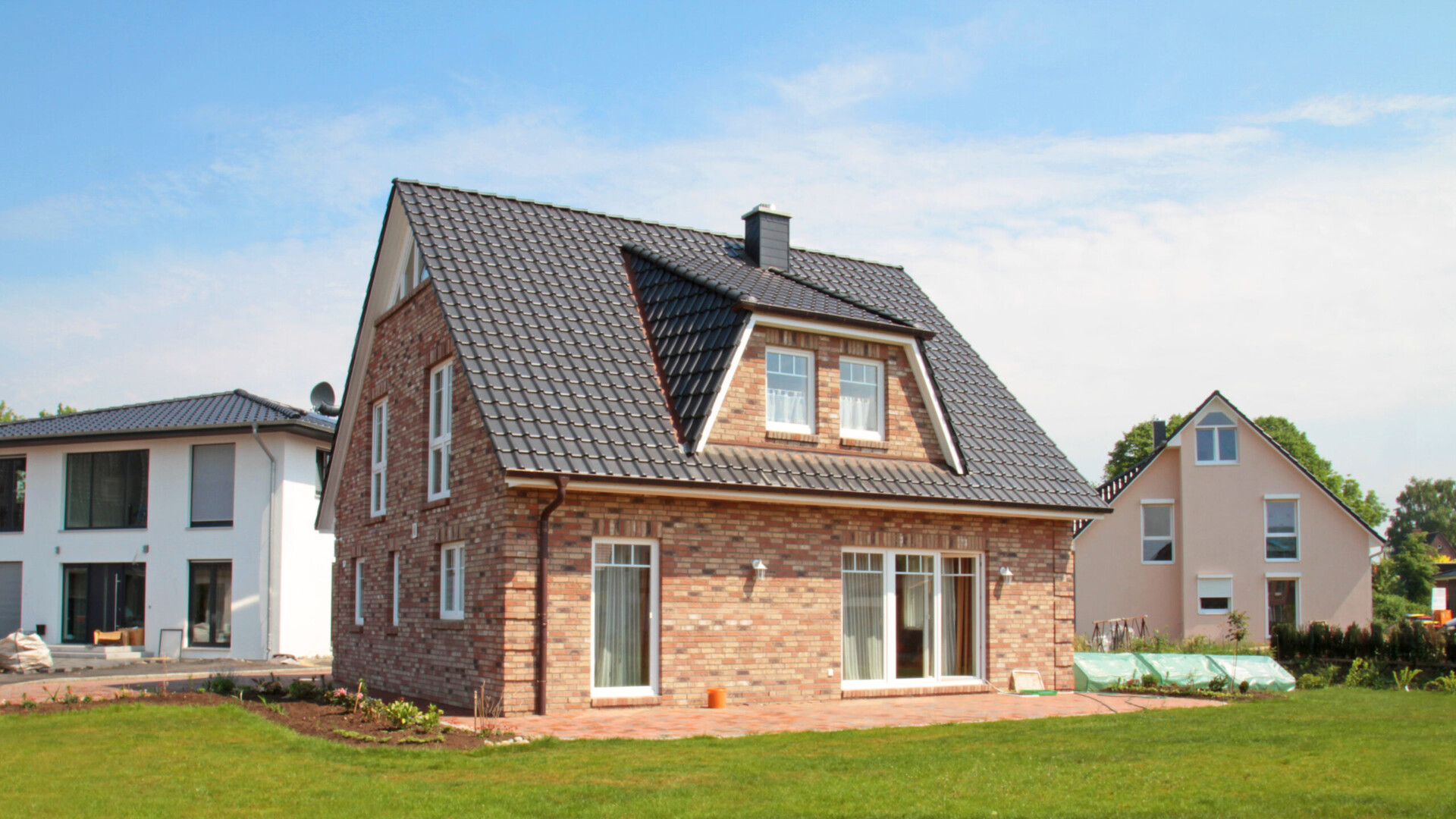 Rotes Backsteinhaus mit dunklem Dach und Dachgaube, umgeben von Rasen und anderen Häusern.