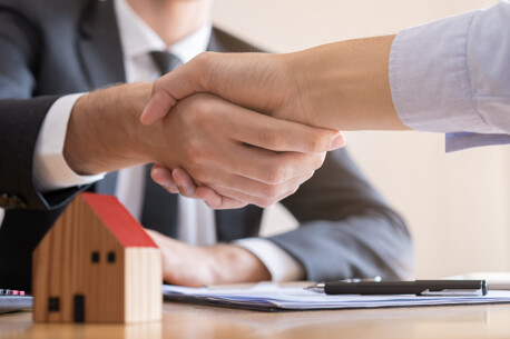 Zwei Personen schütteln sich die Hände über einem Tisch, auf dem ein kleines Holzhausmodell und ein Stift liegen, symbolisiert einen erfolgreichen Geschäftsabschluss im Immobilienbereich.