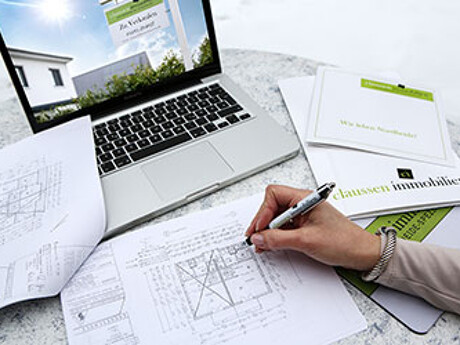 Person arbeitet an Bauplänen neben Laptop mit geöffneter Webseite von 'claussen immobilien' und Briefumschlägen der Firma