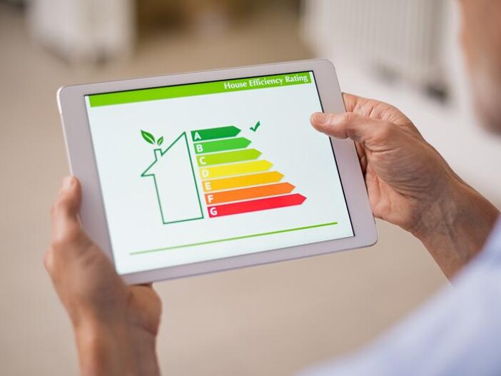 Nahaufnahme eines Mannes, der ein Tablet hält, das einen "House Efficiency Rating"-Bildschirm mit einem Energiesparkonzept und einer Skala von A bis G anzeigt.