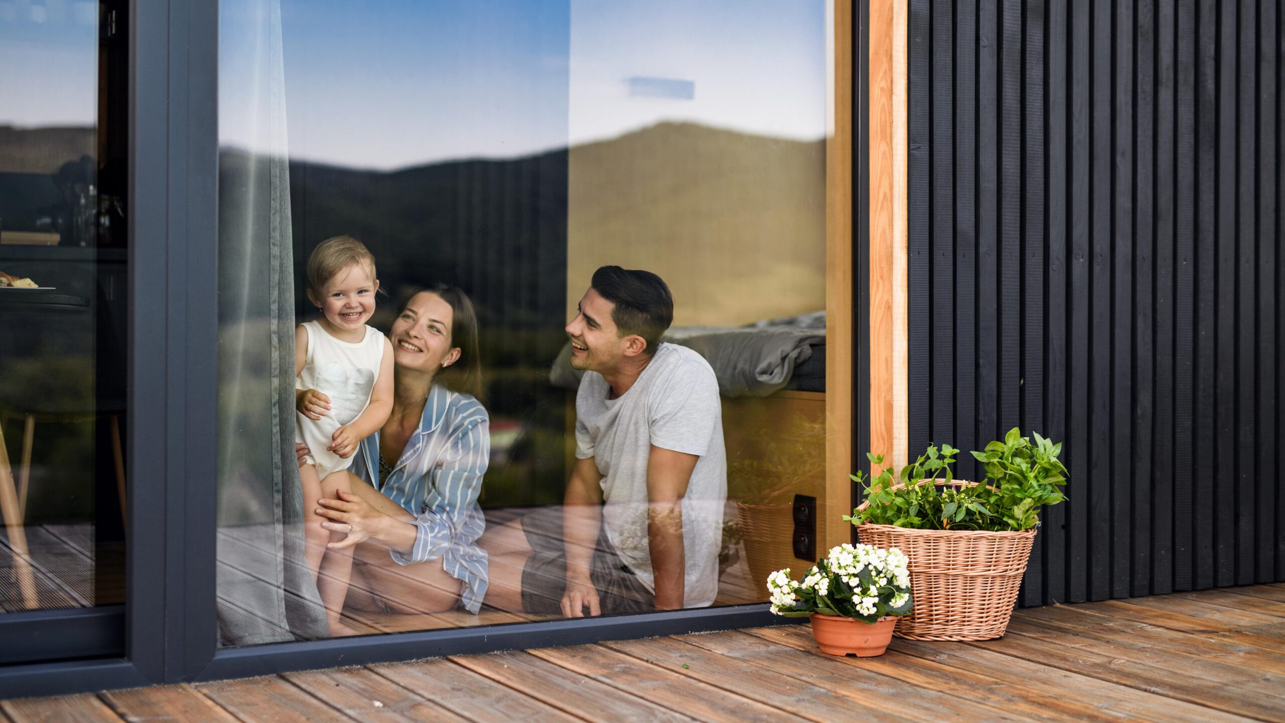 Junge Familie schaut lächelnd durch eine große Glasfront, draußen stehen Pflanzen.
