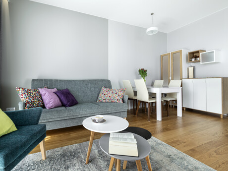 Wohnzimmer mit grauem Sofa, bunten Kissen, blauem Sessel, zwei runden Couchtischen und weißem Esstisch mit vier Stühlen
