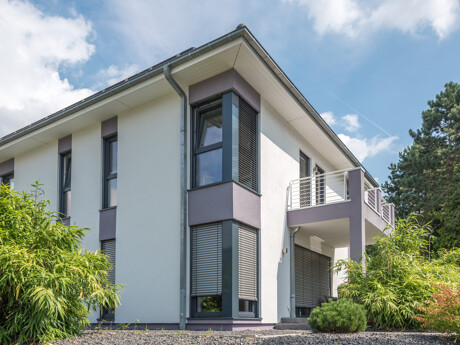 Modernes zweistöckiges Wohnhaus mit weißen Wänden, großen Fenstern und Balkon, umgeben von grünen Pflanzen