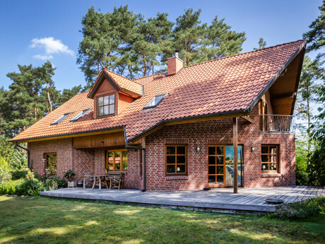 Zweistöckiges Backsteinhaus mit rotem Ziegeldach, mehreren Fenstern und einer Holzterrasse, umgeben von Bäumen und Rasen