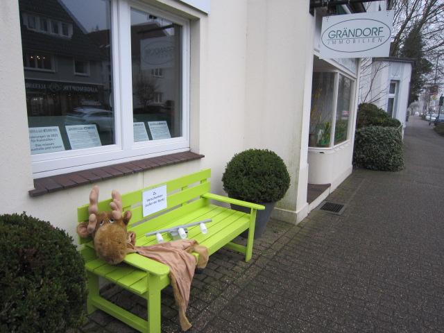 Plüschrentier schlummert auf grüner Bank vor Immobilienbüro.