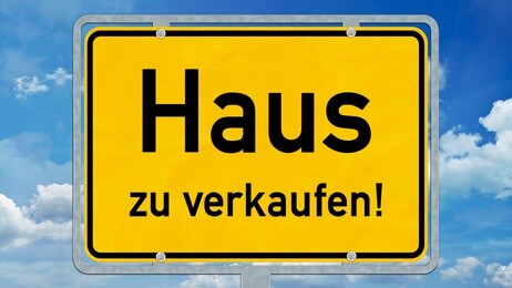 Gelbes Schild mit "Haus zu verkaufen!" vor blauem Himmel.