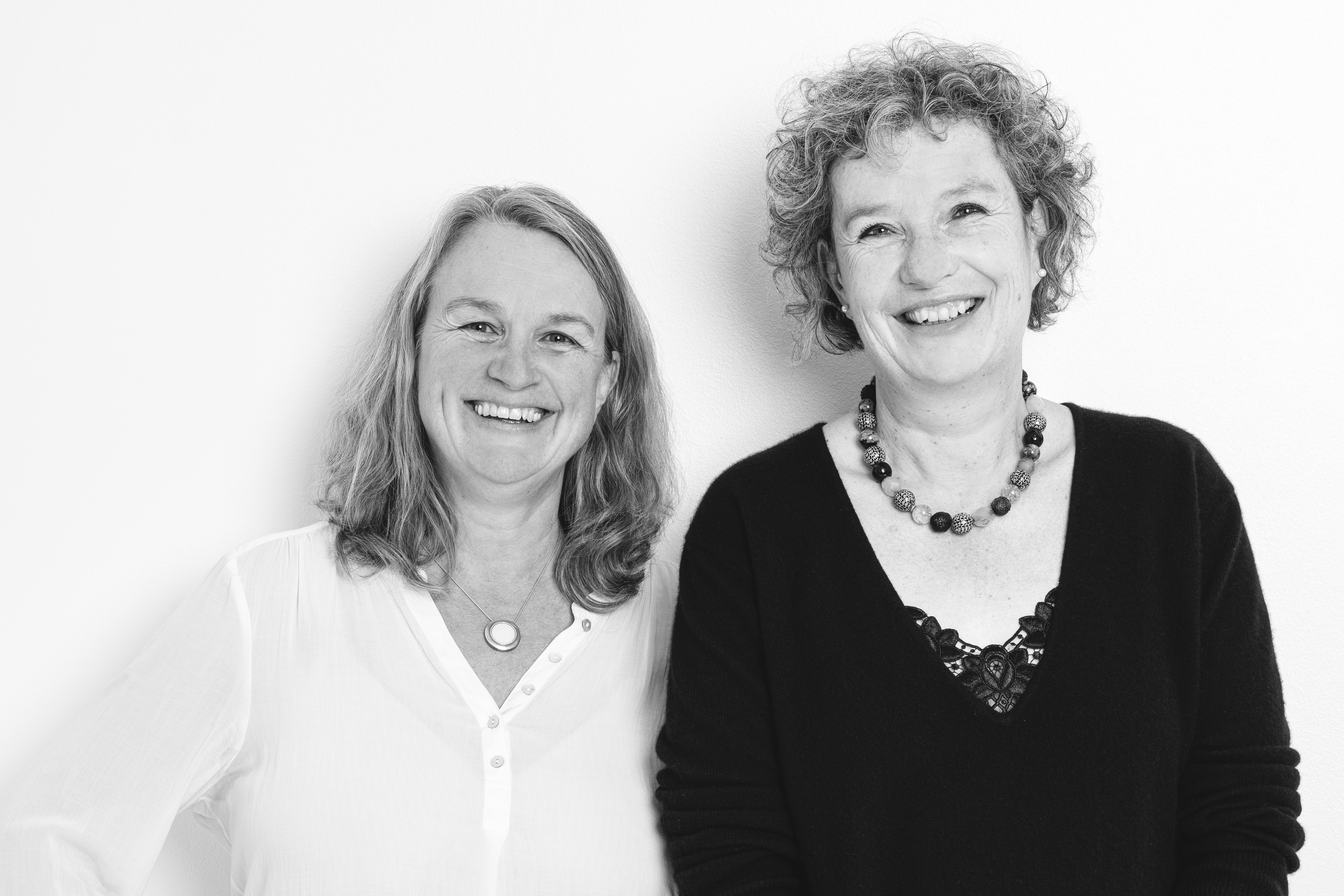 Zwei lächelnde Frauen stehen entspannt vor neutralem Hintergrund.