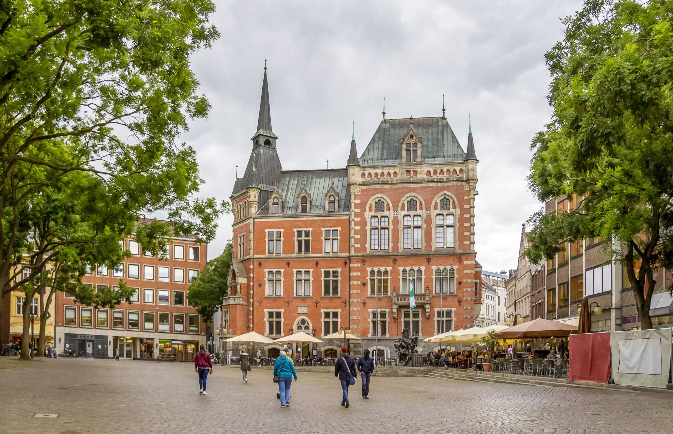 Menschen auf Kopfsteinpflasterplatz vor Backsteingebäude und Cafés.