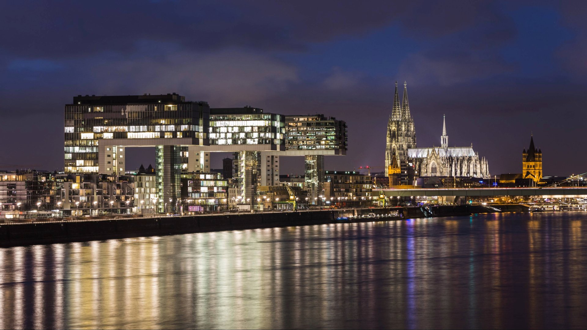 Moderne Kranhäuser am Rheinufer in Köln bei Nacht mit beleuchtetem Dom im Hintergrund.