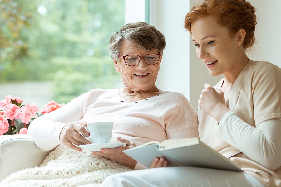 Pflegekraft und Seniorin beim gemeinsamen Lesen in einem hellen Wohnraum auf der Couch.
