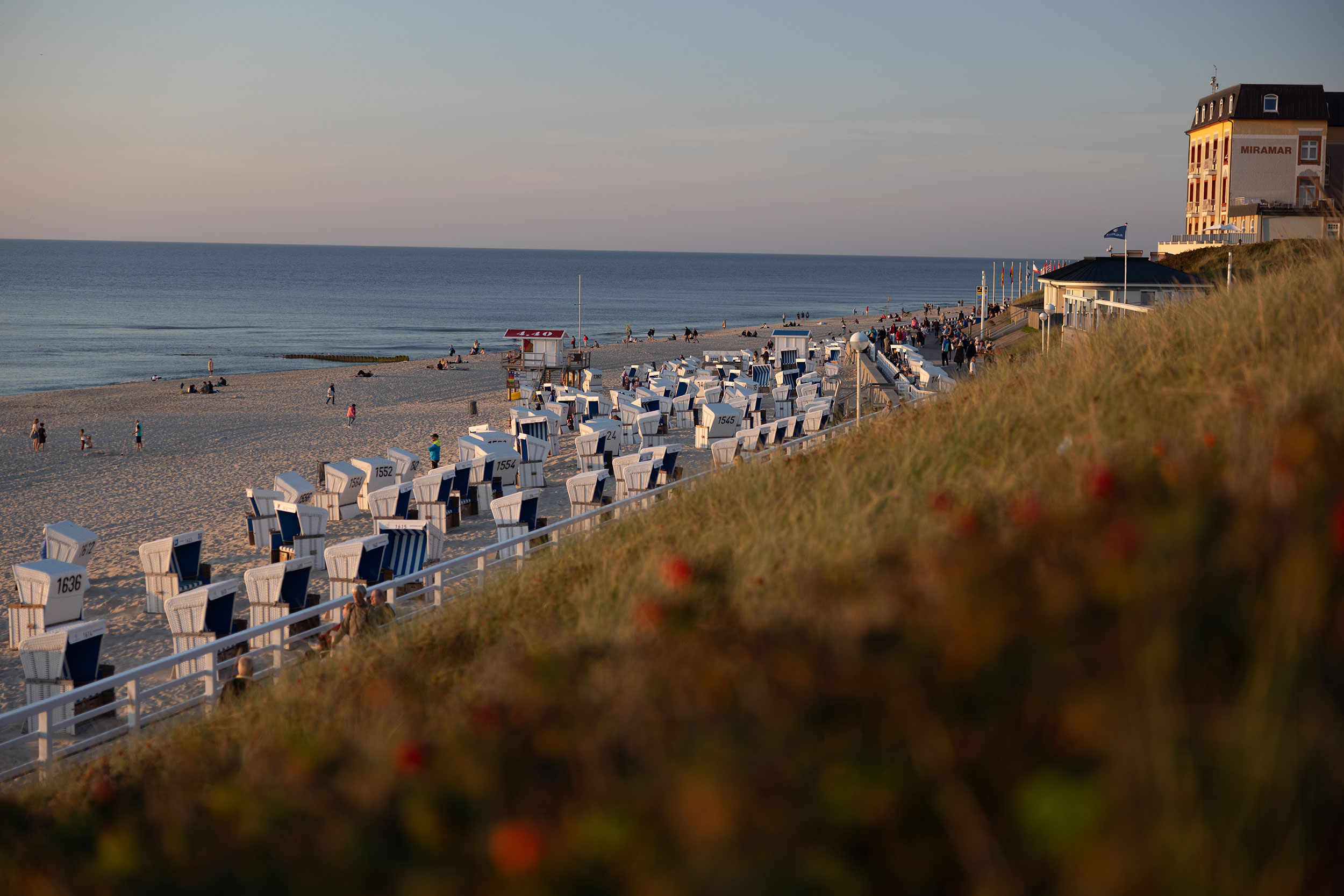 Blick auf einen Sandstrand mit Reihen von weißen Liegestühlen, Menschen, die am Ufer spazieren gehen, und ein großes Gebäude in der Nähe der Küste bei Sonnenuntergang.