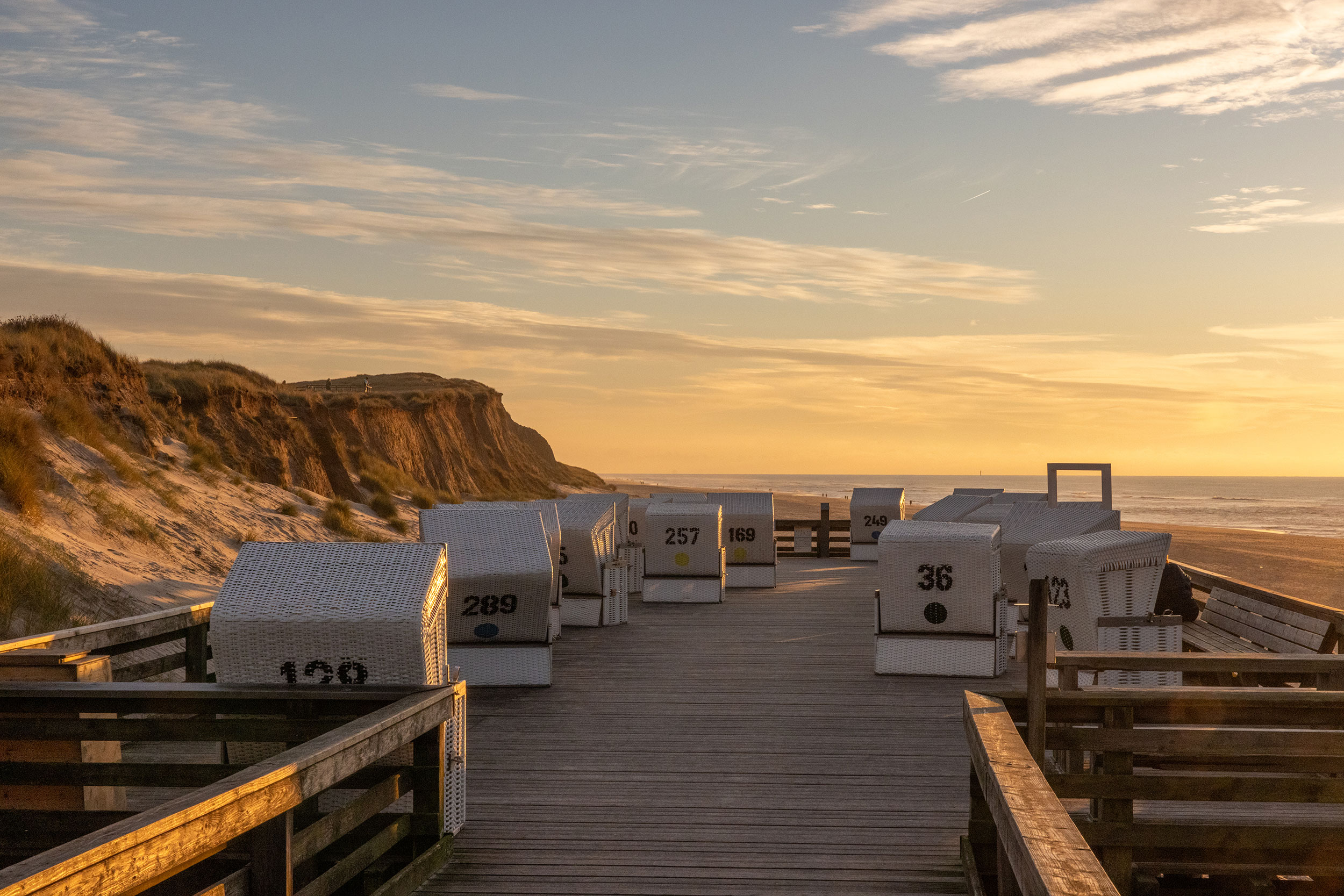 Bei Sonnenuntergang Blick auf eine Holzpromenade mit weißen Strandkörben, nummeriert, mit Blick auf Sanddünen und das Meer.